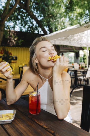 A woman at an outdoor barbecue enjoys a plate of crispy fries and a refreshing drink. Sunlight filters through the trees, creating a relaxed and joyful atmosphere