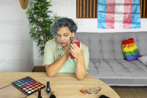 A young transgender man thoughtfully applies makeup, sitting at a table with various makeup items. A pride flag is visible in the background, symbolizing LGBTQ+ identity