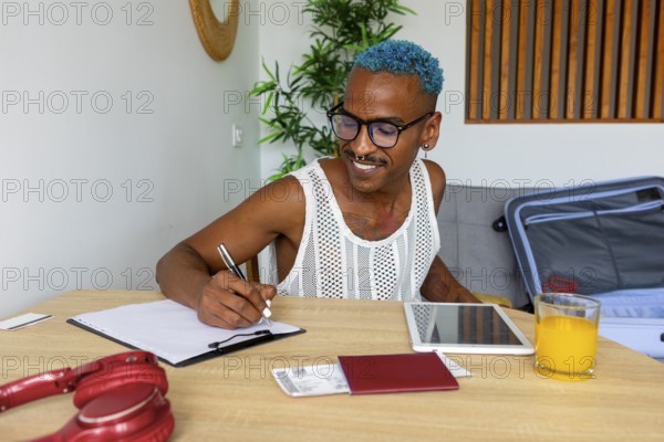 An LGBTQIA+ man enthusiastically prepares for holidays, jotting down plans at a table. Notebooks, a tablet, and a suitcase are ready for the upcoming adventure