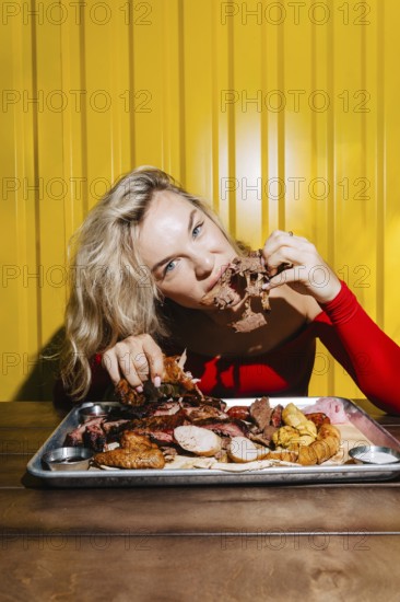 A woman savors a delicious barbecue platter filled with an assortment of meats against a bright yellow backdrop. The scene captures the joy of dining and the vibrant atmosphere