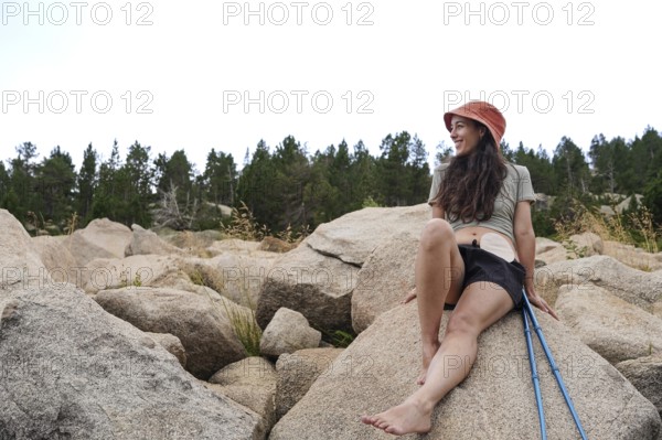 A smiling woman with an ostomy enjoys nature, sitting on large rocks amidst a serene forest backdrop She embraces outdoor life, embodying strength and positivity