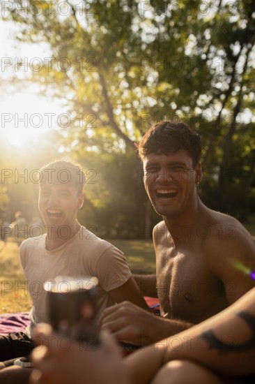 Two young men sit laughing by a lake during summer, one holding a drink. Warm sunlight illuminates their joyful expressions