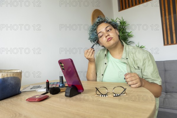 Transgender man applying makeup while recording a tutorial with a smartphone placed on a stand, sitting at a wooden table with makeup items and glasses