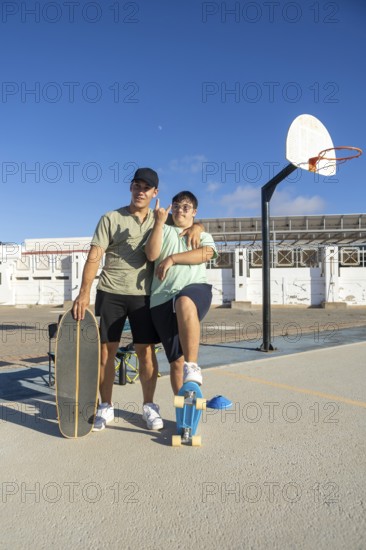 A teenager with Down Syndrome poses confidently with his brother and skateboards on an outdoor court, expressing pride, inclusion, and joy in a sunny and vibrant setting
