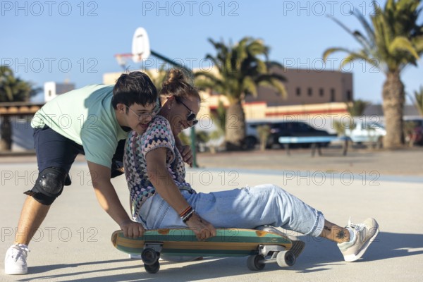 A teenager with Down syndrome helps his mother ride a skateboard outdoors, sharing a fun and energetic moment together in a sunny and relaxed environment
