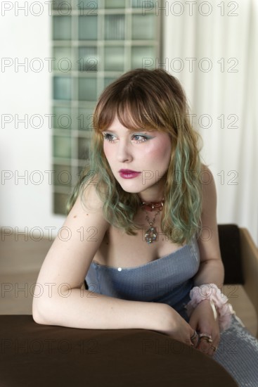 A woman with colorful, pastel hair leans thoughtfully on a brown table Dressed in a light blue outfit with accessories, she gazes into the distance, framed by soft curtains and glass tiles