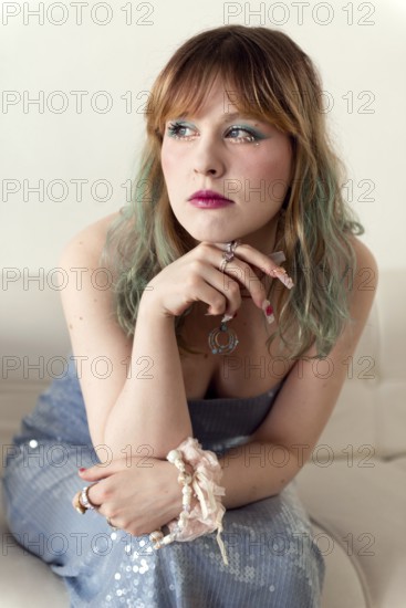 A woman with green-tinted hair and expressive makeup is seated on a white couch She wears a blue sequined dress, numerous rings, and a fabric bracelet, gazing pensively
