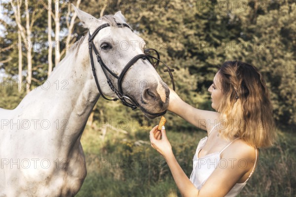 A young woman feeds a white horse a carrot in a peaceful forest. The tranquil scene captures nature, companionship, and gentle interaction between human and animal