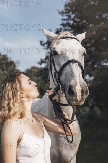 A young woman in a white dress shares a serene moment with a white horse outdoors. The sunlight highlights their bond, enhancing the peaceful and natural setting