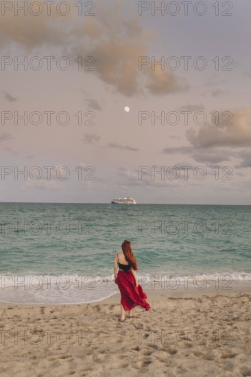 Back view of an unrecognizable redhead woman strolling along Miami Beach. She wears a flowing red skirt and looks at the ocean with a cruise ship in the distance under a twilight sky