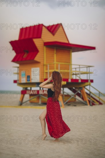 Back view of unrecognizable redhead woman walking near a vividly colored lifeguard station on Miami Beach, capturing a tranquil layover moment