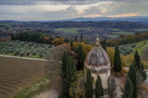 Aerial view of the Chapel of San Michele Arcangelo in Semifonte, Tuscany, surrounded by rolling hills, olive groves, vineyards, and cypress trees under a dramatic sunset sky