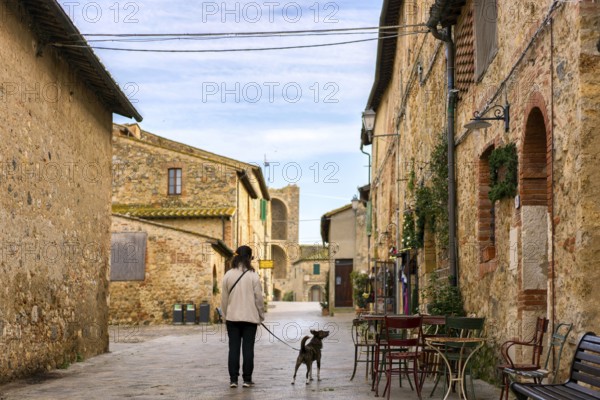 A serene street scene in Monteriggioni, Tuscany, with a woman walking her dog past rustic stone buildings. Outdoor tables and medieval architecture create a timeless village atmosphere