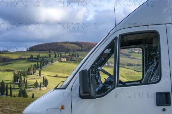 A camper van parked in the scenic Tuscan countryside, surrounded by rolling hills, cypress trees, and picturesque farmland under a cloudy sky, capturing tranquil rural vibes