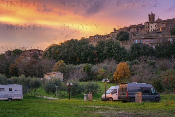 Camper vans and motorhomes parked in a scenic service area below a traditional Tuscany village. The hilltop stone houses and vibrant sunset sky create a picturesque countryside setting