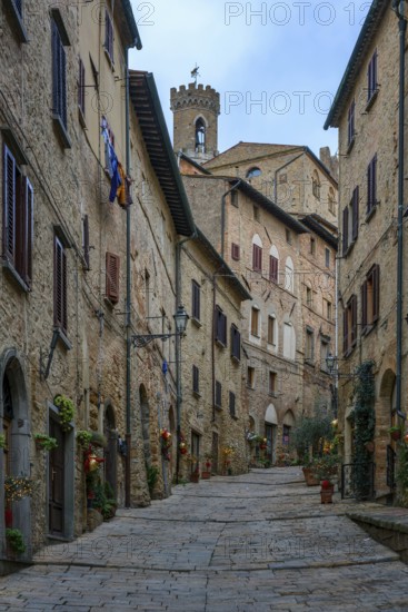 A charming cobblestone street in Volterra, Tuscany, lined with traditional stone houses adorned with flowers. The medieval architecture and tower create a timeless Italian village atmosphere