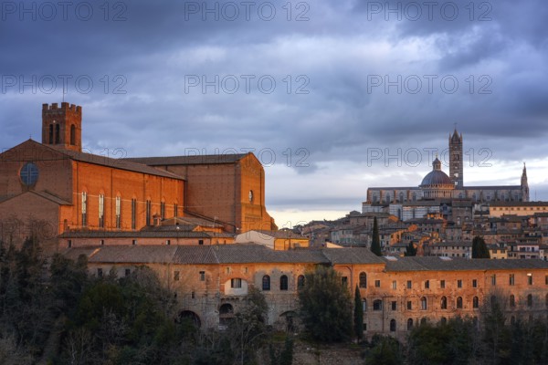 A stunning view of Siena at sunset, featuring the illuminated San Domenico Basilica and the Siena Cathedral. The warm light highlights the medieval architecture against a dramatic cloudy sky