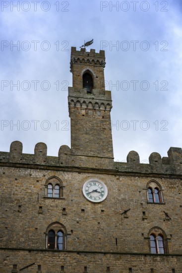 A close-up of the Priori Palace tower in Volterra, Tuscany, showcasing its medieval architecture and iconic clock. The stone structure stands tall against a cloudy sky, embodying Italian heritage
