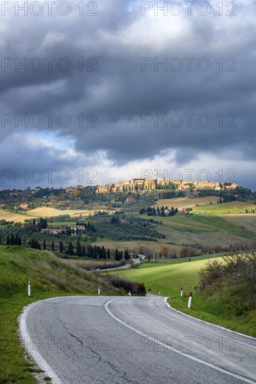 A scenic view of Pienza from the main road in Val d Orcia, Tuscany. The winding road cuts through rolling green hills under a dramatic cloudy sky, highlighting the charm of the Italian countryside