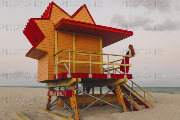 A redhead woman enjoys a quiet moment on the stairs of an iconic orange and red lifeguard station during a sunset at Miami Beach, embodying a relaxed and scenic layover
