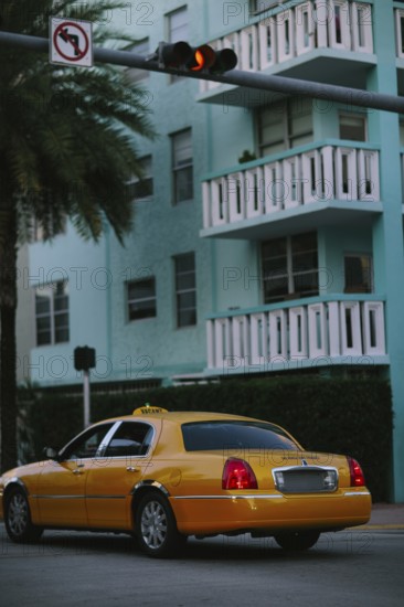A bright yellow taxi cab parked on a palm-lined street in Miami with a multi-story turquoise building in the background, depicting urban transportation