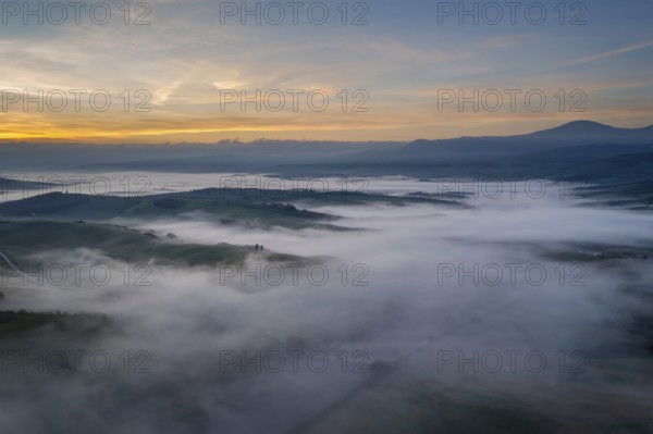 A stunning aerial view of the Val d Orcia in Tuscany, Italy, with rolling hills blanketed in morning mist under a colorful sunrise, creating a tranquil and dreamy landscape