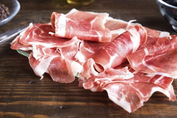 Thin, delicately rolled slices of fresh ham displayed on a dark wooden surface, highlighted by natural lighting that enhances the rich, pink hues and marbled texture of the meat