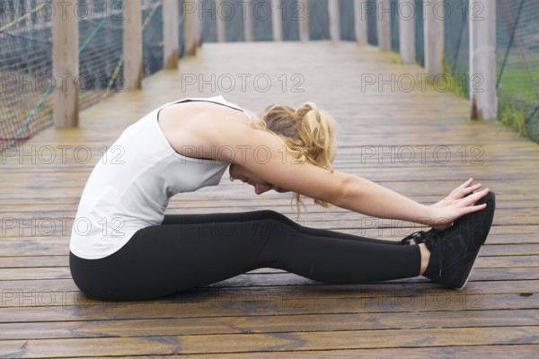 Young blonde woman wearing sportswear sits stretches her legs on a wooden bridge, focusing on her exercise routine