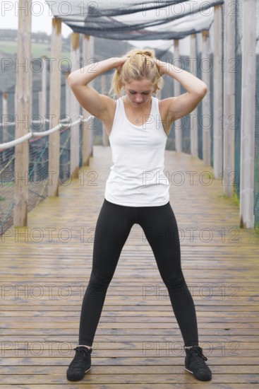 A young blond woman exercises by stretching outdoors on a rustic wooden deck, dressed in sporty black leggings and a white tank top, focused and determined in her fitness routine