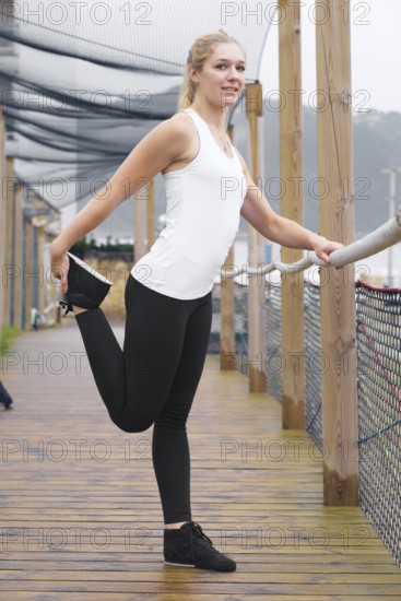 A young blond woman, dressed in athletic wear, stretches on a wooden walkway, displaying a healthy lifestyle and sportsmanship