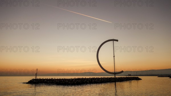 The serene scene of a sunset at Lake Geneva, showcasing a large ring-like sculpture standing on a jetty, silhouetted against a vibrant sky