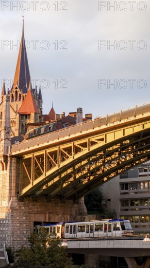 A metro train crosses a modern bridge in the foreground with the historic Lausanne Cathedral rising majestically in the background. The warm glow of sunset illuminates the scene