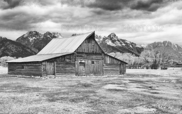 A black and white image capturing an old wooden barn with the majestic Grand Teton range in the background under a cloudy sky. Mormon Row in Wyoming, USA
