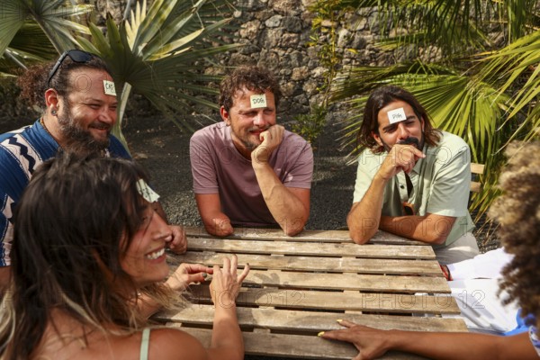 A multiethnic group of friends plays a guessing game at a wooden table outdoors They laugh while trying to identify the words on sticky notes on their foreheads, enjoying a sunny day