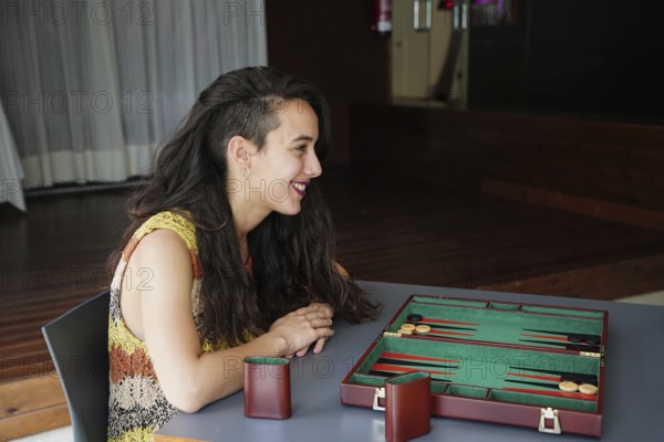 A woman with long dark hair smiles as she sits across a backgammon board in a cozy indoor setting The board is set up for a strategic game, suggesting relaxation and leisure