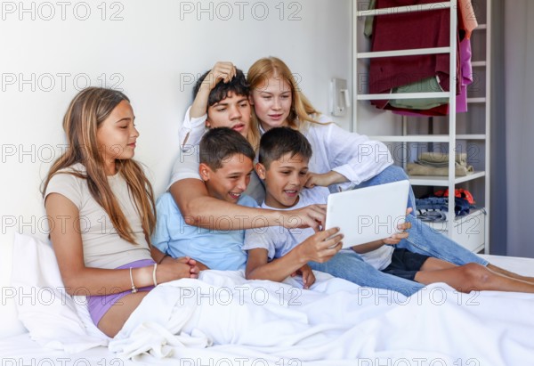 Five siblings, including a young teen woman, a young man, and triplet kids (a girl and two boys), share a joyful family time exploring a tablet together while relaxing on a bed at their home
