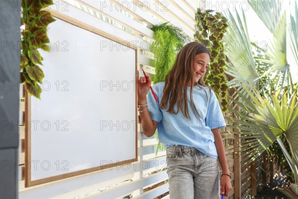 A cheerful young girl stands next to a whiteboard surrounded by lush tropical foliage, holding a marker and enjoying a sunny day outdoors