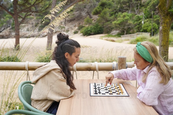 Mother and daughter entertain themselves in a lively game of chess on a wooden table Surrounded by vegetation and sand, they concentrate intensely on each move, combining strategy with nature