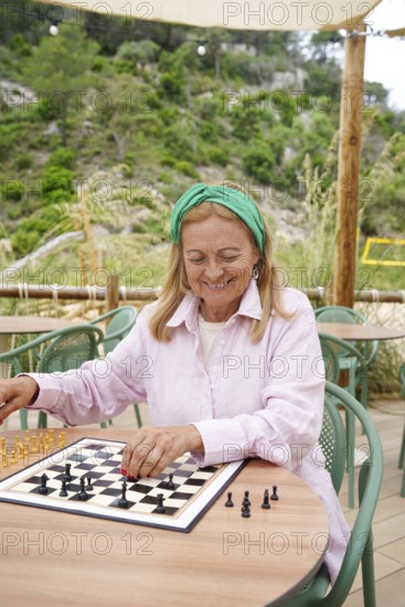 A senior woman plays chess outdoors on a sunny day, seated at a round table surrounded by greenery She smiles while contemplating her next move, embracing relaxation