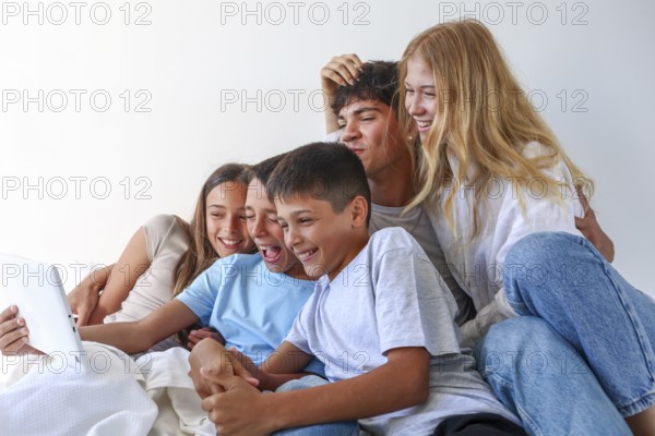 A joyful moment at home with five siblings, an older brother and sister with their younger triplet siblings, consisting of a girl and two boys, all enjoying and laughing over a tablet