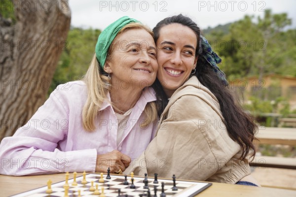 Mother and daughter enjoy a sunny day playing chess outside, sharing a joyful bonding moment The chessboard sits on a wooden table amidst a lush, green environment