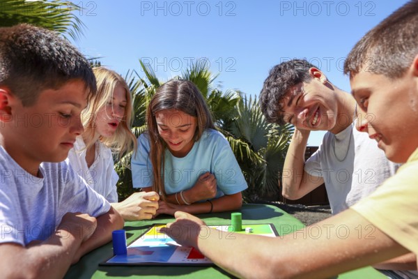 Two older siblings, a young man and woman, enjoy a board game outside with their younger triplets siblings, a girl and two boys, on a sunny day, promoting family bonding and fun