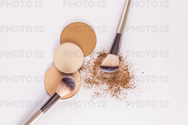 Top view of various shades of compact powders next to a makeup brush, with loose powder scattered on a white background