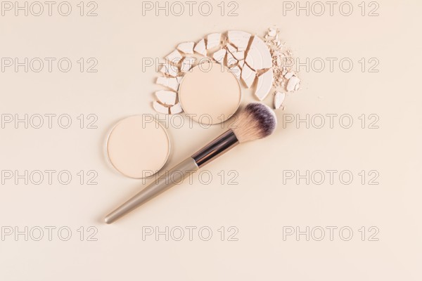 Top view of a styled makeup layout featuring compact powder in whole and broken forms, and loose powder next to a makeup brush on a pastel backdrop