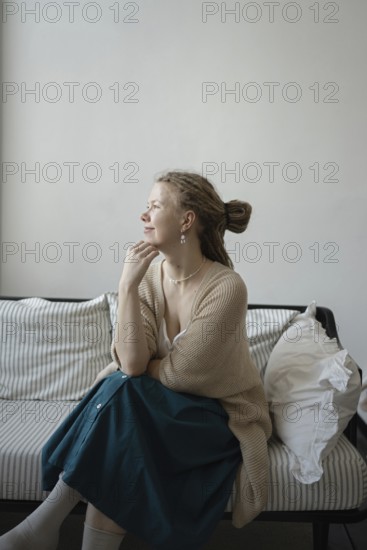 A serene woman sits thoughtfully on a striped sofa, wearing a cozy cardigan and skirt The calm atmosphere is enhanced by soft lighting and minimalist decor, evoking tranquility