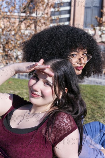 Two Gen Z friends, one Hispanic girl with curly hair and another girl with straight black hair, sharing a playful moment outdoors. The girl with curly hair is looking away from the camera while shielding her eyes from the sun, displaying a joyful expression