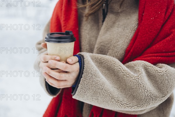 Cropped unrecognizable young woman in a red coat and cozy scarf holding a warm coffee cup outdoors on a snowy winter day