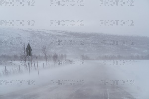 A blizzard envelops a remote road in Swedish Lapland, obscuring distant snow-covered hills and roadside fences. The winter landscape is stark and atmospheric