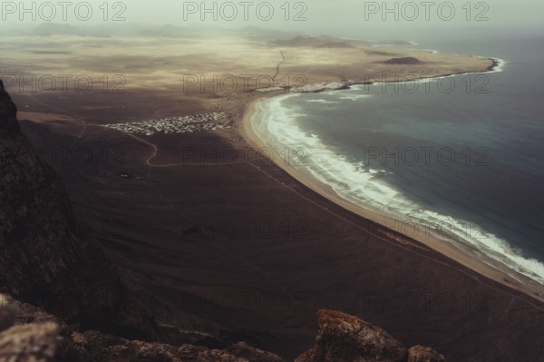 Stunning aerial view of Lanzarote's rugged coastline, showcasing a sweeping sandy beach meeting the turquoise ocean, enveloped by dramatic volcanic terrain and distant mountains