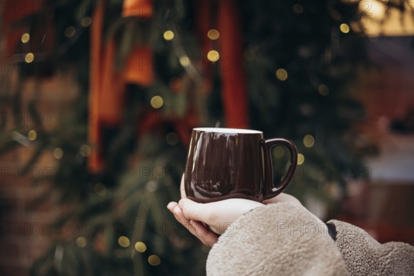 Cropped unrecognizable young woman holding a warm coffee mug in front of a beautifully decorated Christmas tree, capturing the essence of comfort and warmth during the winter season